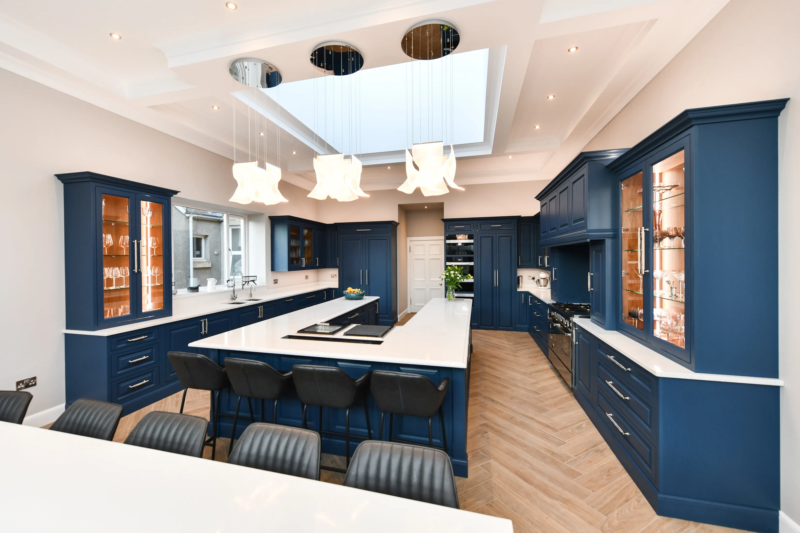 Large handmade kitchen featuring a large U shaped central island, lots of storage, lit glass cabinets and a pantry, all viewed from the large seating area